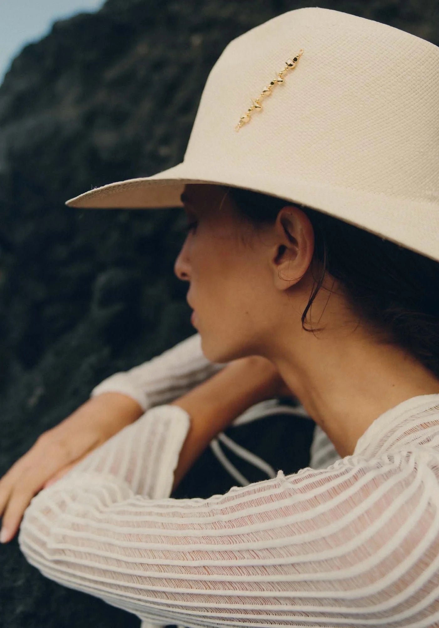 La femme est appuyée sur un rocher et porte le chapeau de paille Diana avec broche dorée natural de chez Van Palma