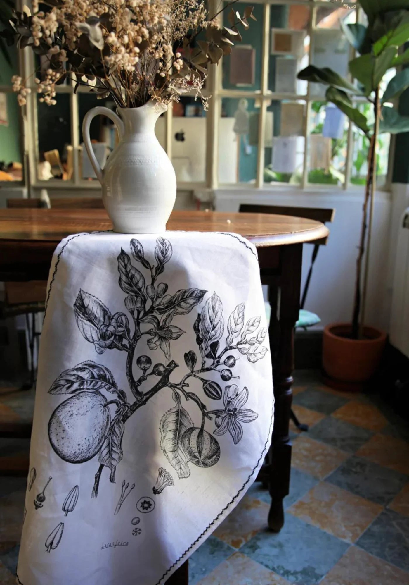 Le linge de cuisine et tenture murale Laranjeira de chez Luz suspendu au bord d'une table et retenu par un vase.