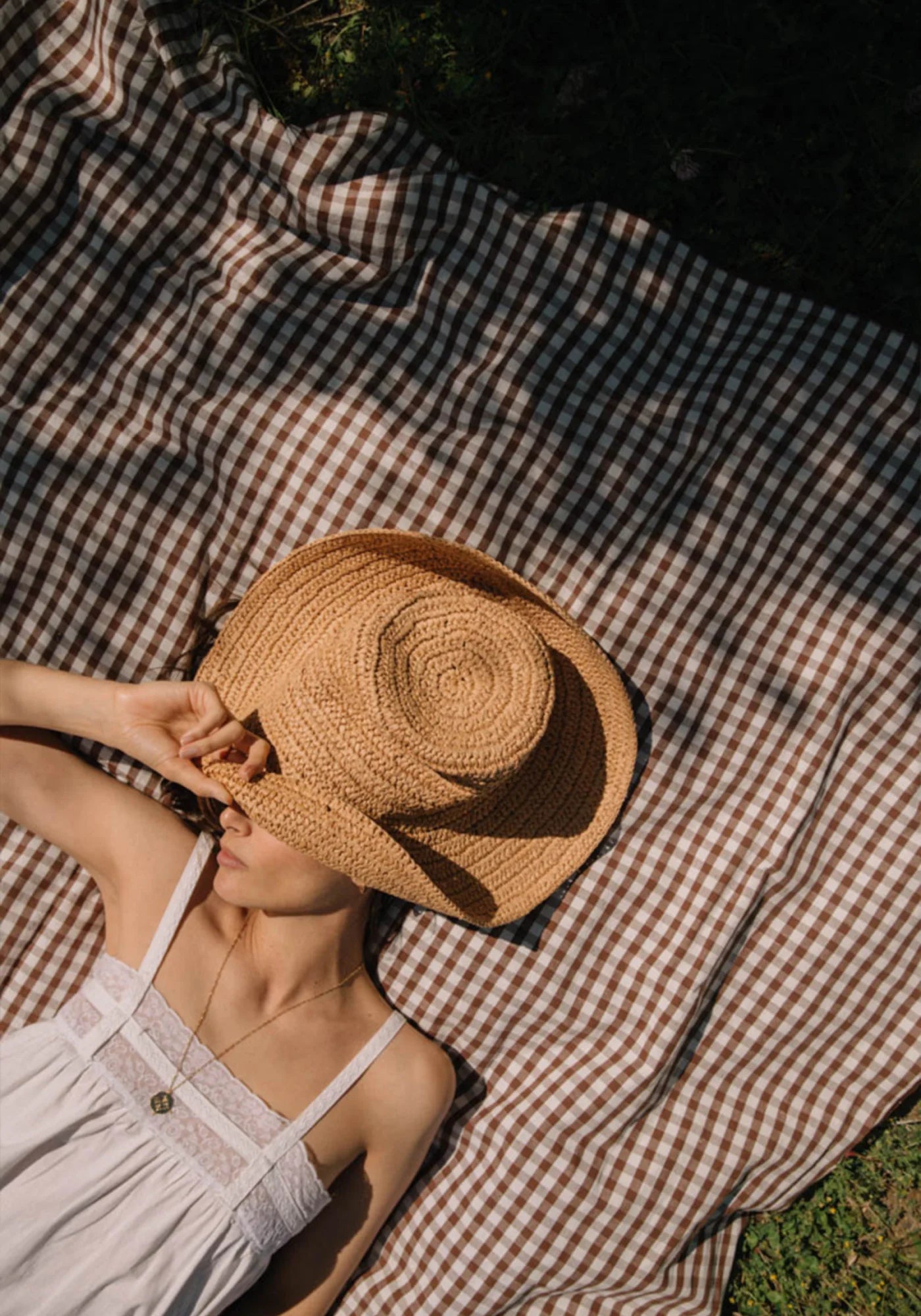 La femme est allongée sur la nappe en lin et coton vichy noisette de chez Les Pensionnaires en cachant son visage avec un chapeau de paille