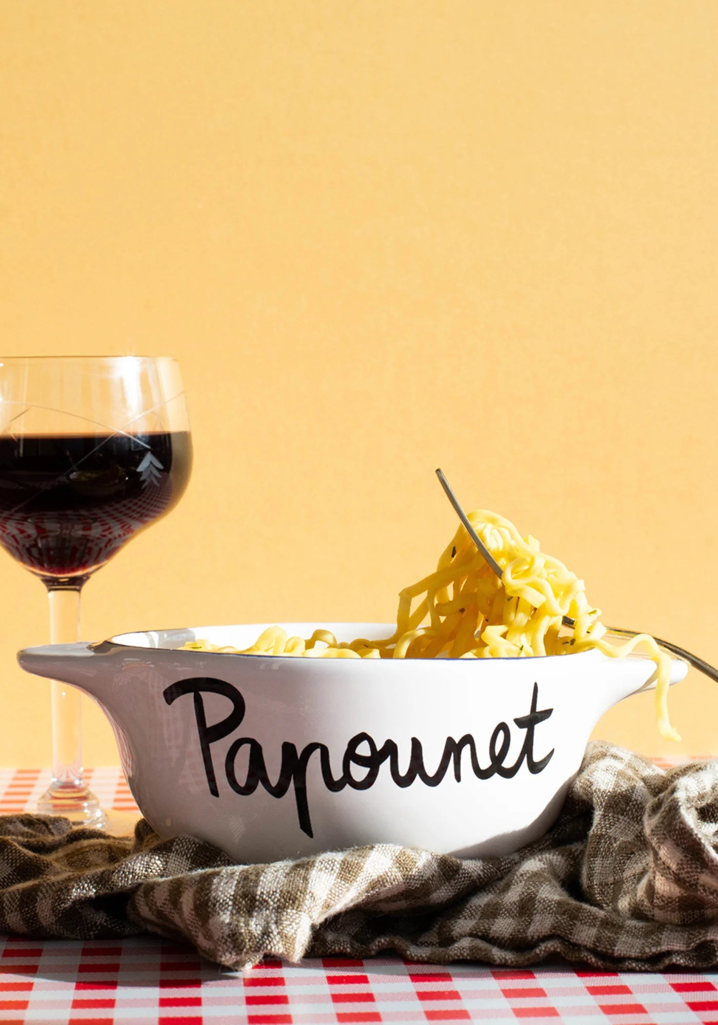 Le bol breton en faïence Papounet de chez Pied de Poule posé sur une nappe à carreaux et un torchon, rempli de spaghettis et accompagné d'un verre de vin rouge