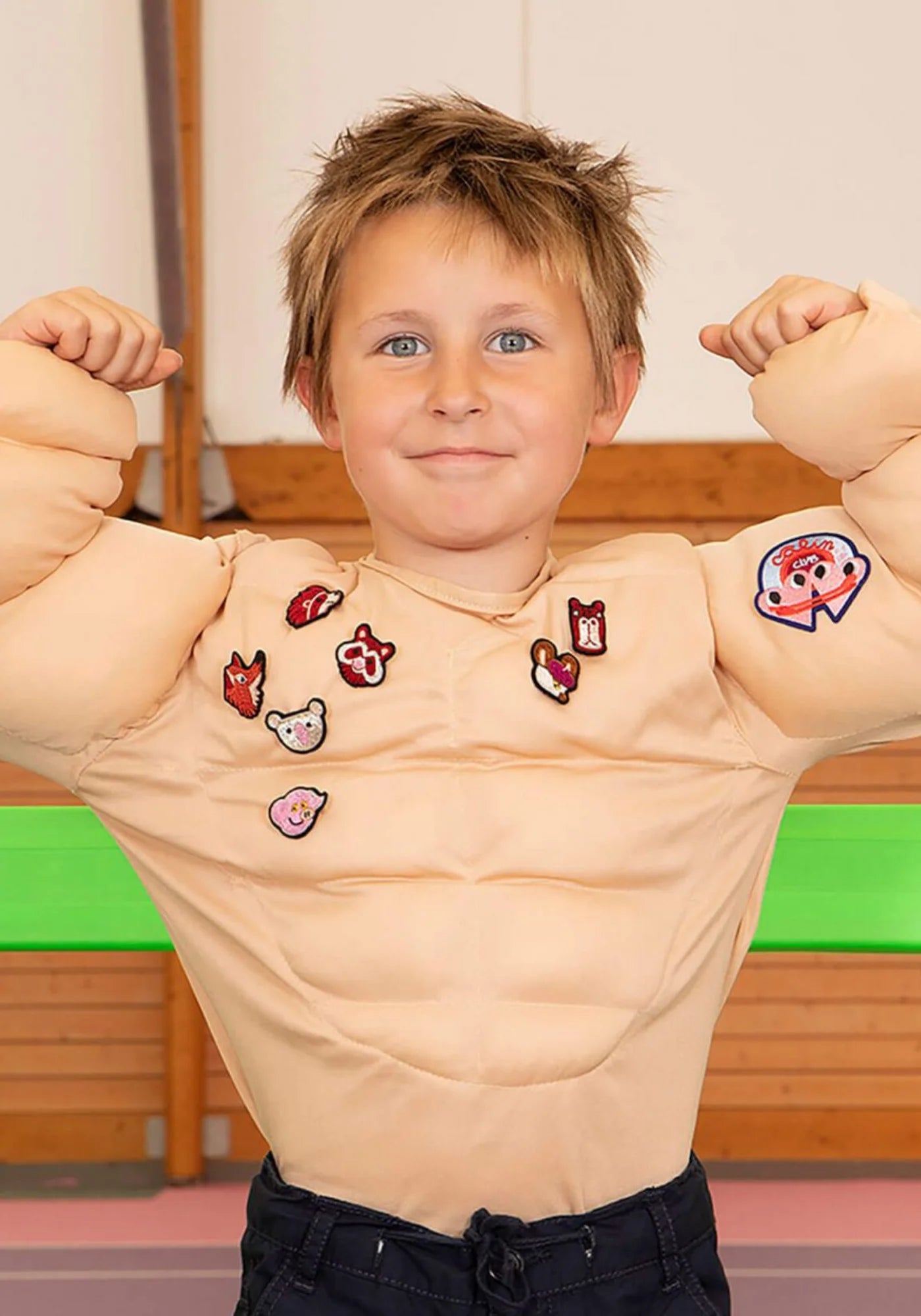 Le petit garçon porte un costume d'un corps musclé en levant les bras pour montrer ses muscles. Sur le costume, la broche brodée à la main Billy Piggy de chez Macon & Lesquoy