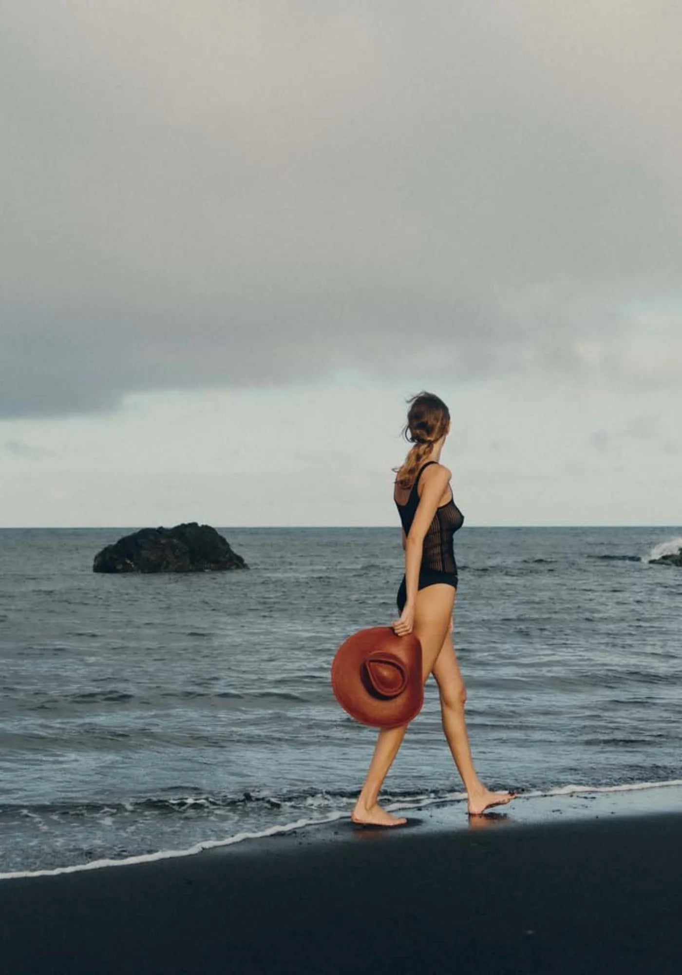 La femme marche sur la plage au bord de l'eau et porte à la main le chapeau de paille Pat auburn de chez Van Palma