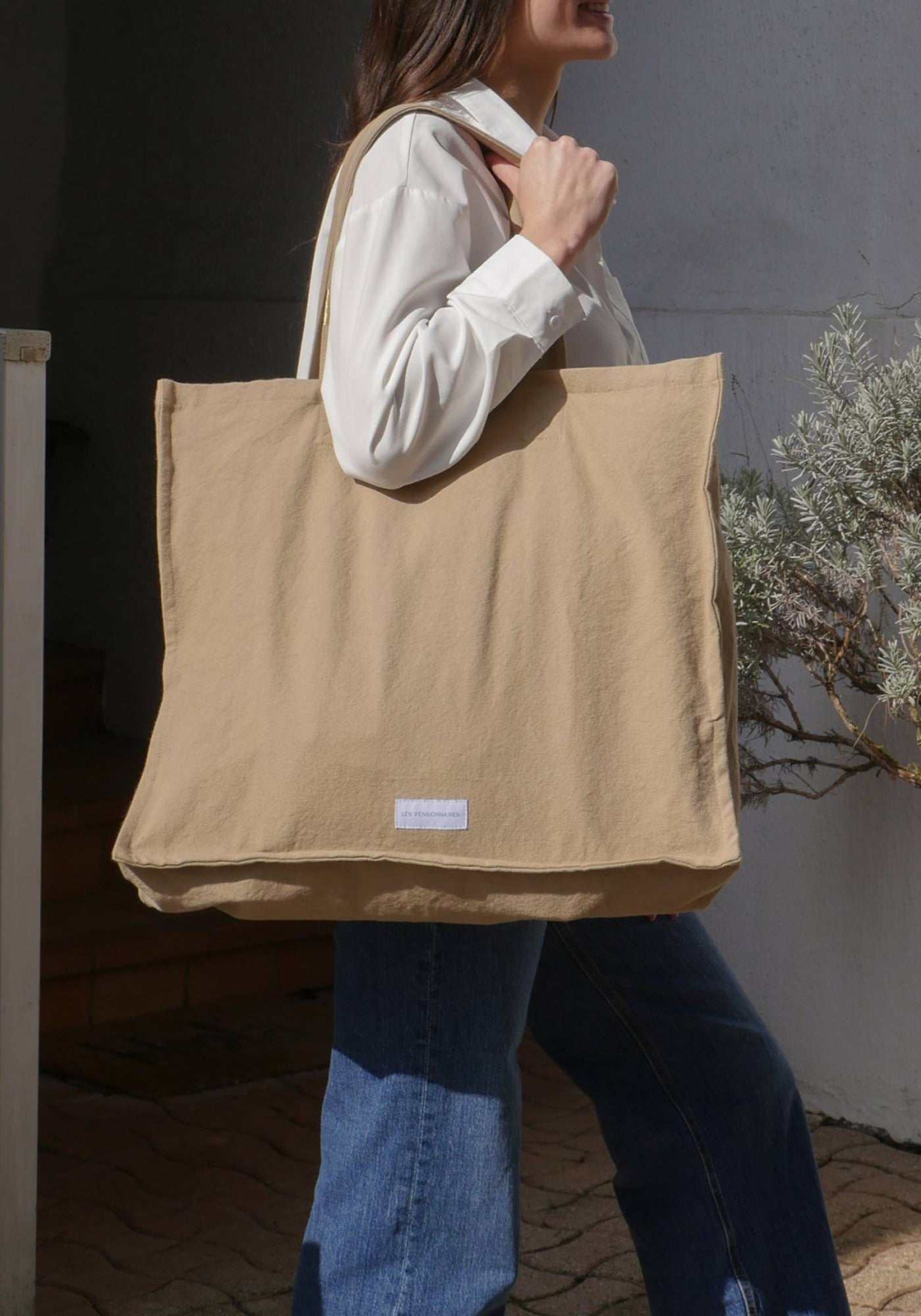 La femme porte sur son épaule le grand sac cabas beige sable de chez Les Pensionnaires