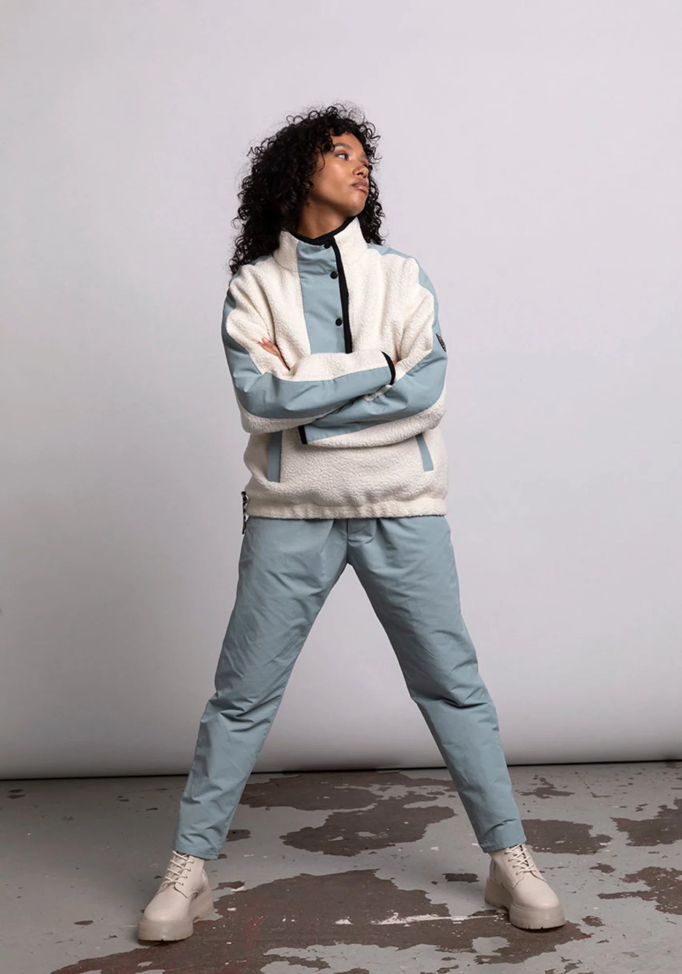 La femme regarde sur le côté avec les bras croisés et les jambes écartées. Elle porte le hoodie Sherpa unisexe Cocoon écrue et bleue de chez Mout Mout