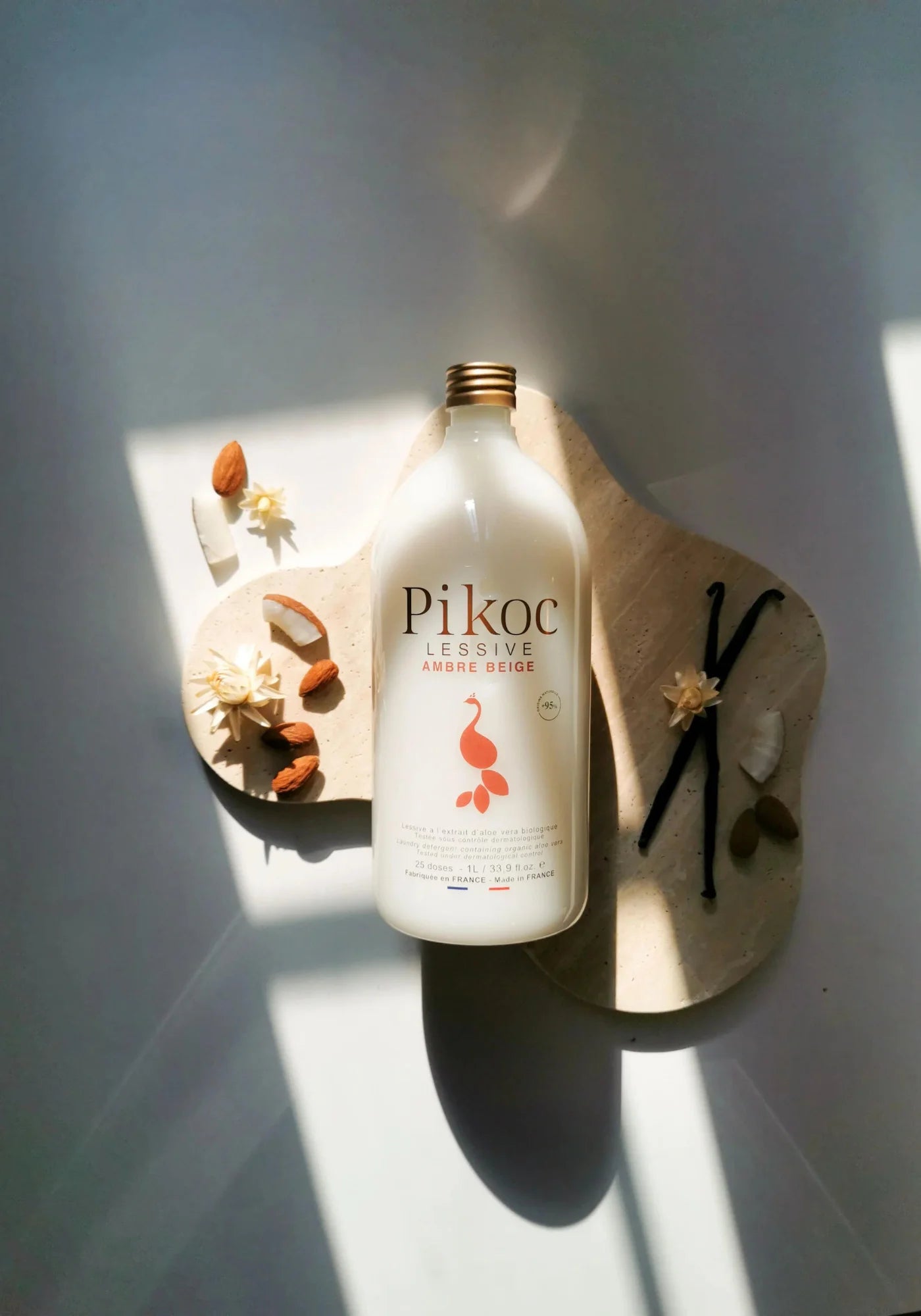 La lessive ambre beige de chez Pikoc posée sur une planche en bois et entourée d'amandes, de gousses de vanille et de noix de coco