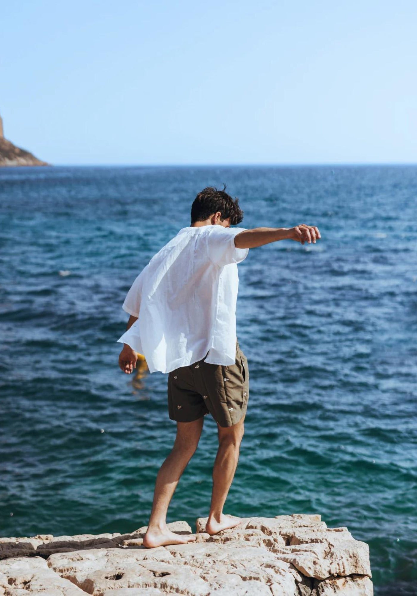 L'homme tient l'équilibre sur un rocher face à la mer et porte le short de bain Les Oliviers de chez Calanque
